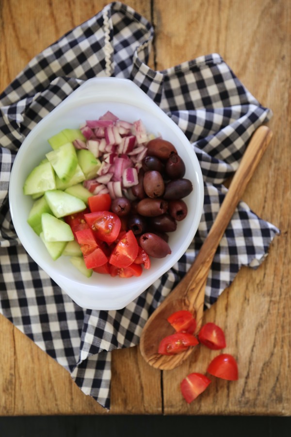 White bowl filled with cucumbers, tomatoes, olives, and onions, with a wooden spoon on the table nearby.