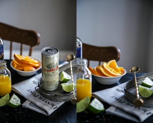 Ginger beer, orange juice, oranges, limes, and a cocktail spoon on a table with a newspaper.