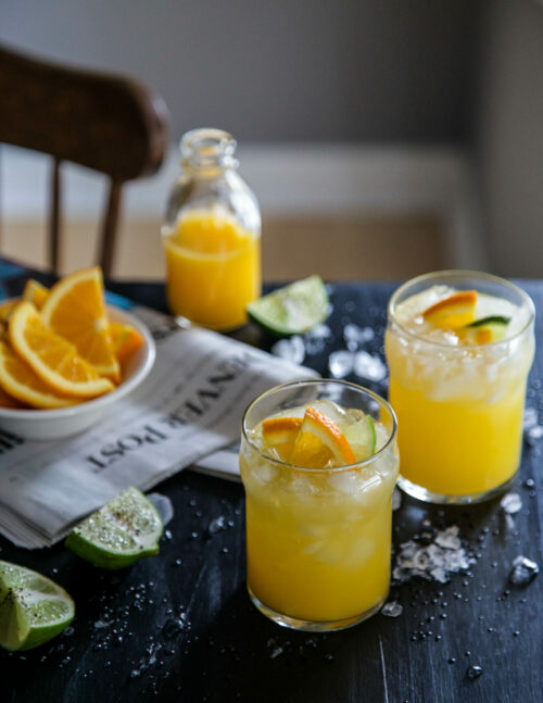 Two morning cocktails with oranges and limes and crushed ice on a table.