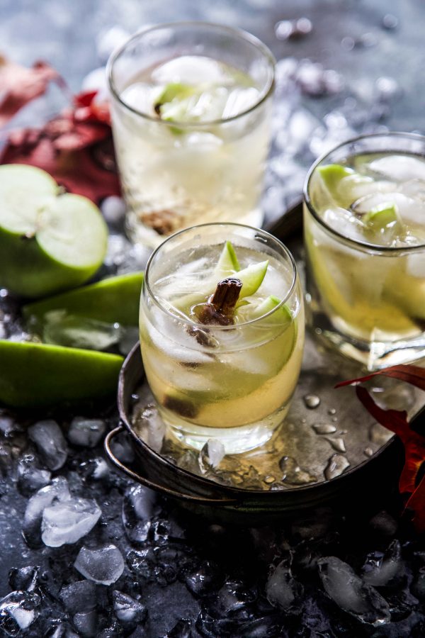 Three glasses of apple ginger cocktail with ice, slices of green apple, and cinnamon sticks.