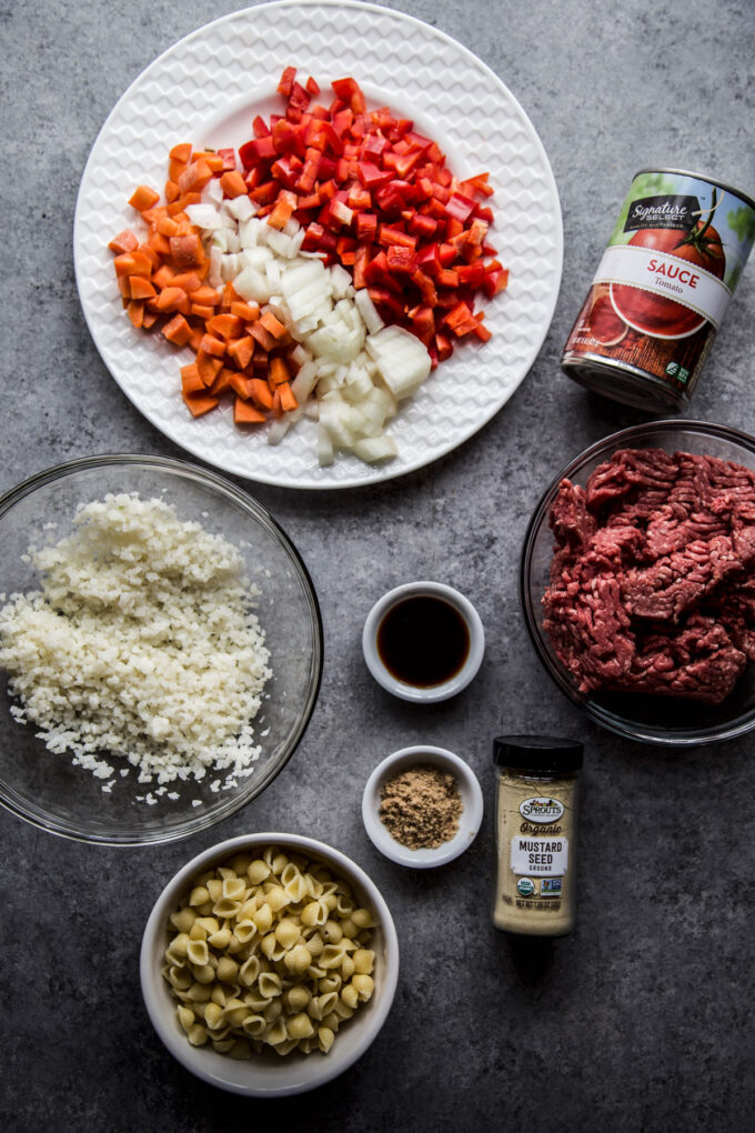 Ingredients in bowls on a table - diced veggies, ground beef, tomato sauce, cauliflower rice, spices, and pasta.